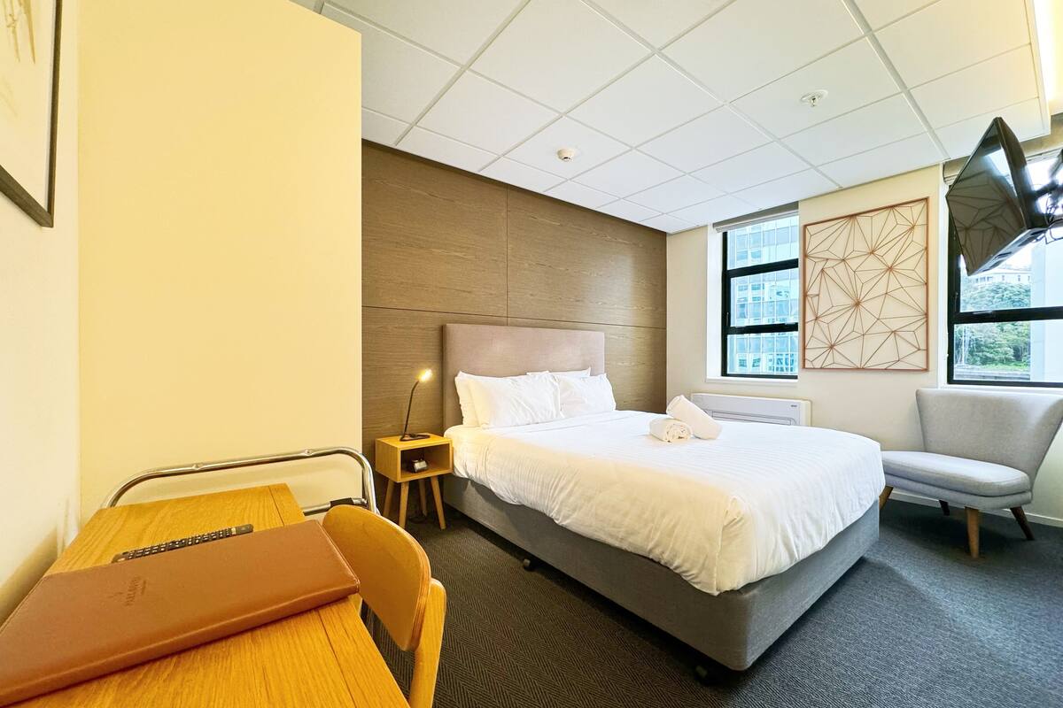 Habitación de hotel con una cama grande, un escritorio con una computadora portátil, una silla, una mesita de noche con una lámpara y una ventana con vistas a edificios.
