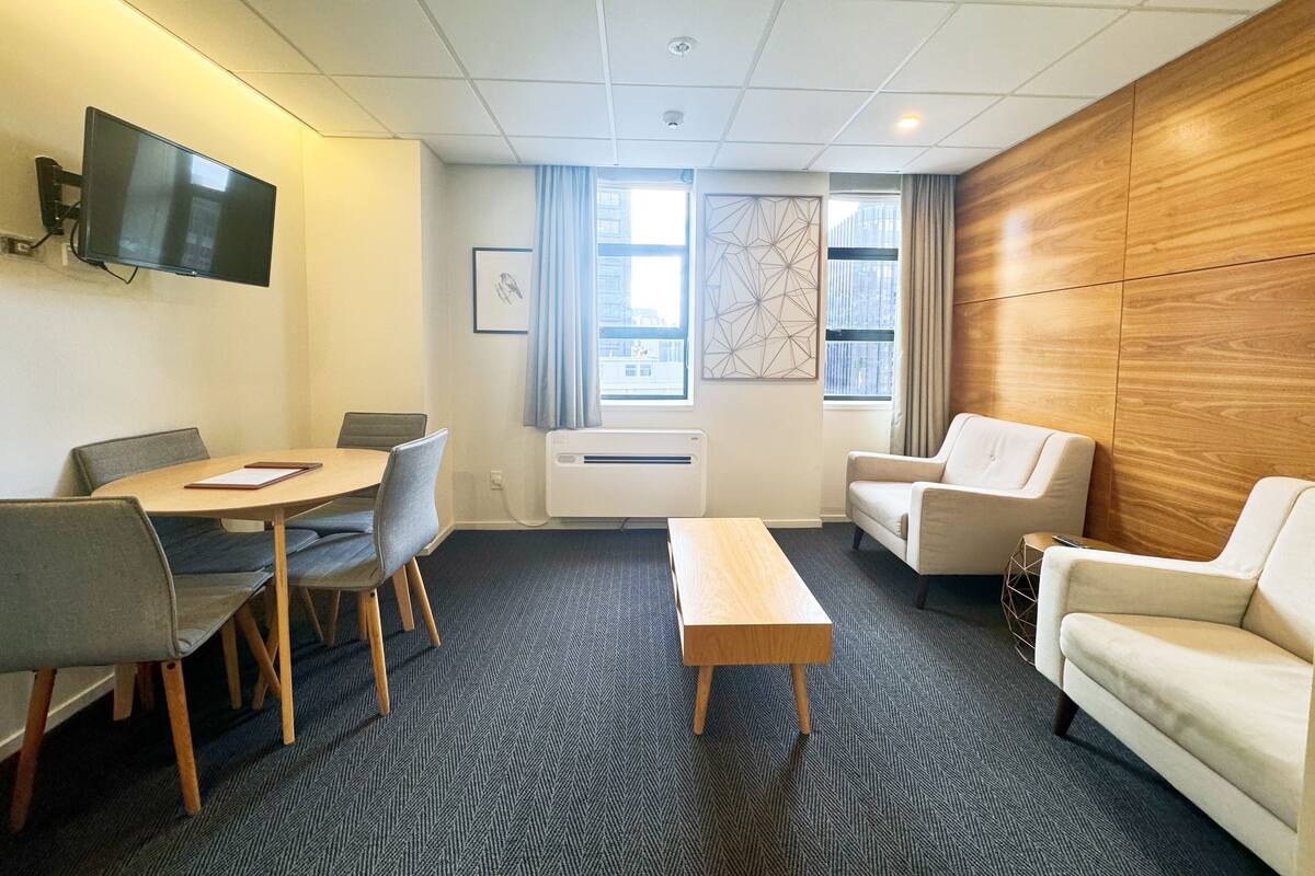 Una habitación de hotel moderna con zona de comedor, sofá, televisor y un amplio ventanal con vistas a la ciudad.