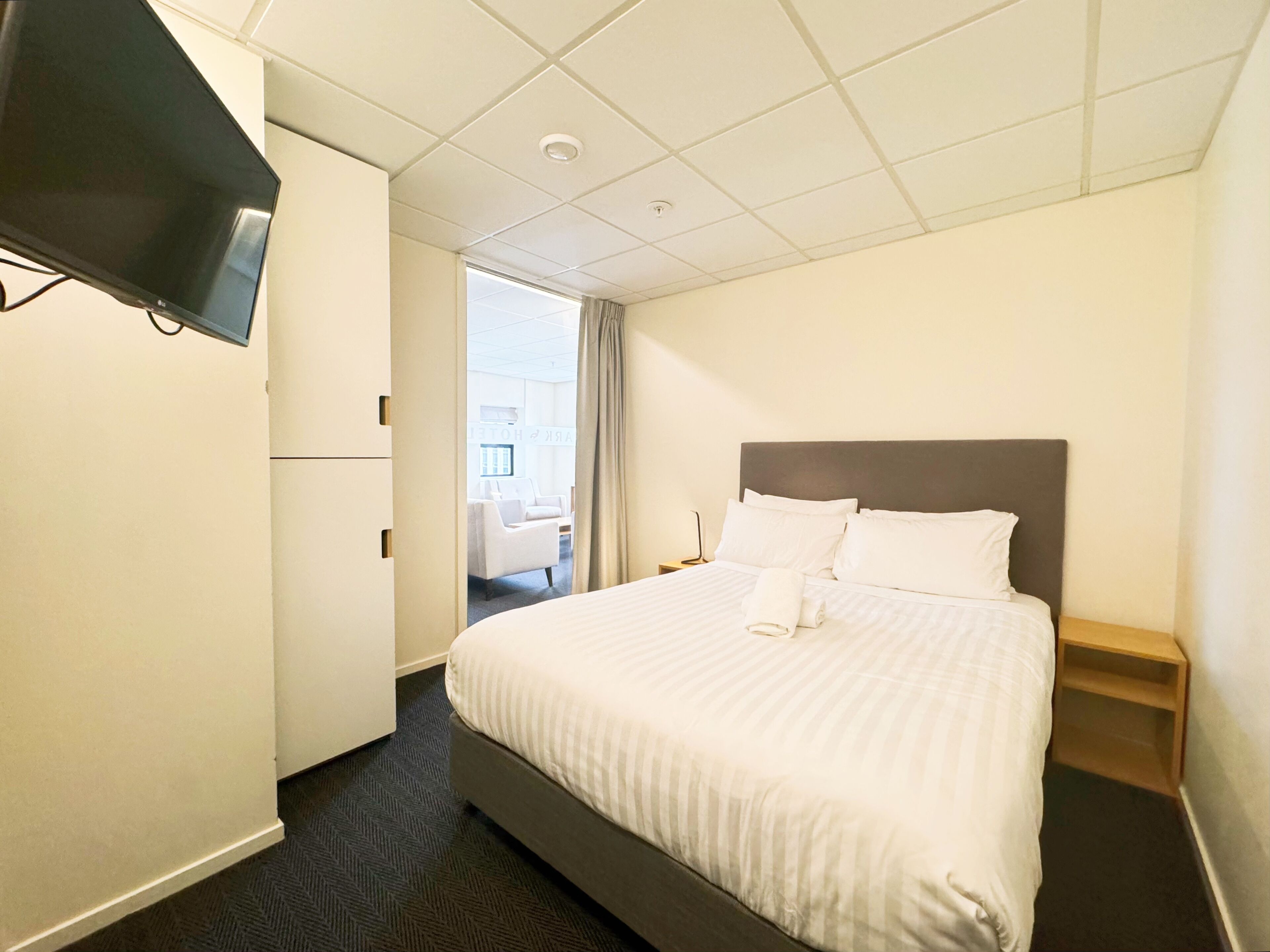 A hotel room with a large bed, a flat-screen TV mounted on the wall, a desk with a chair, and a view of the hotel's lobby.