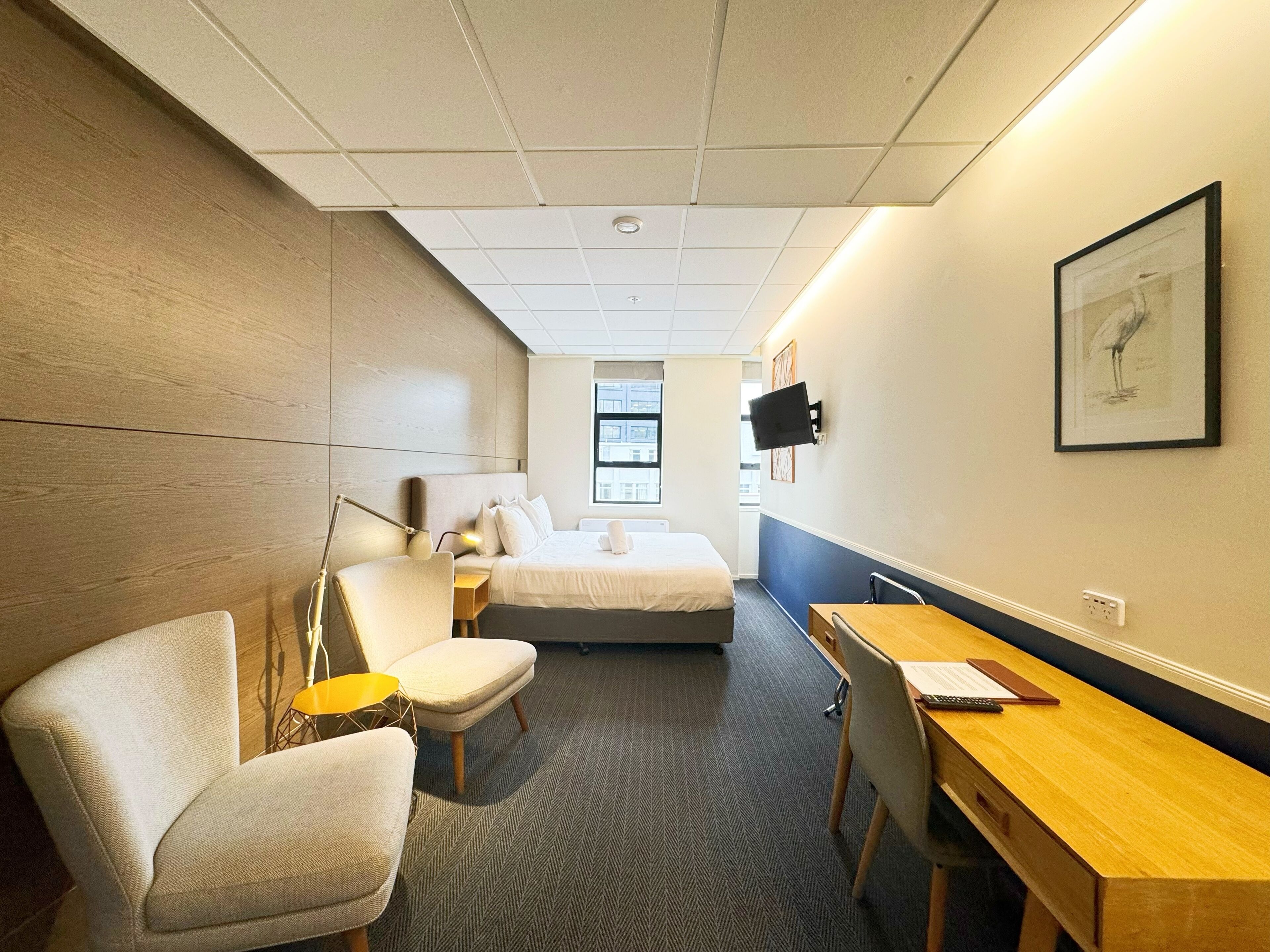 A hotel room with a bed, two chairs, a desk, a lamp, a TV, and a framed picture on the wall.