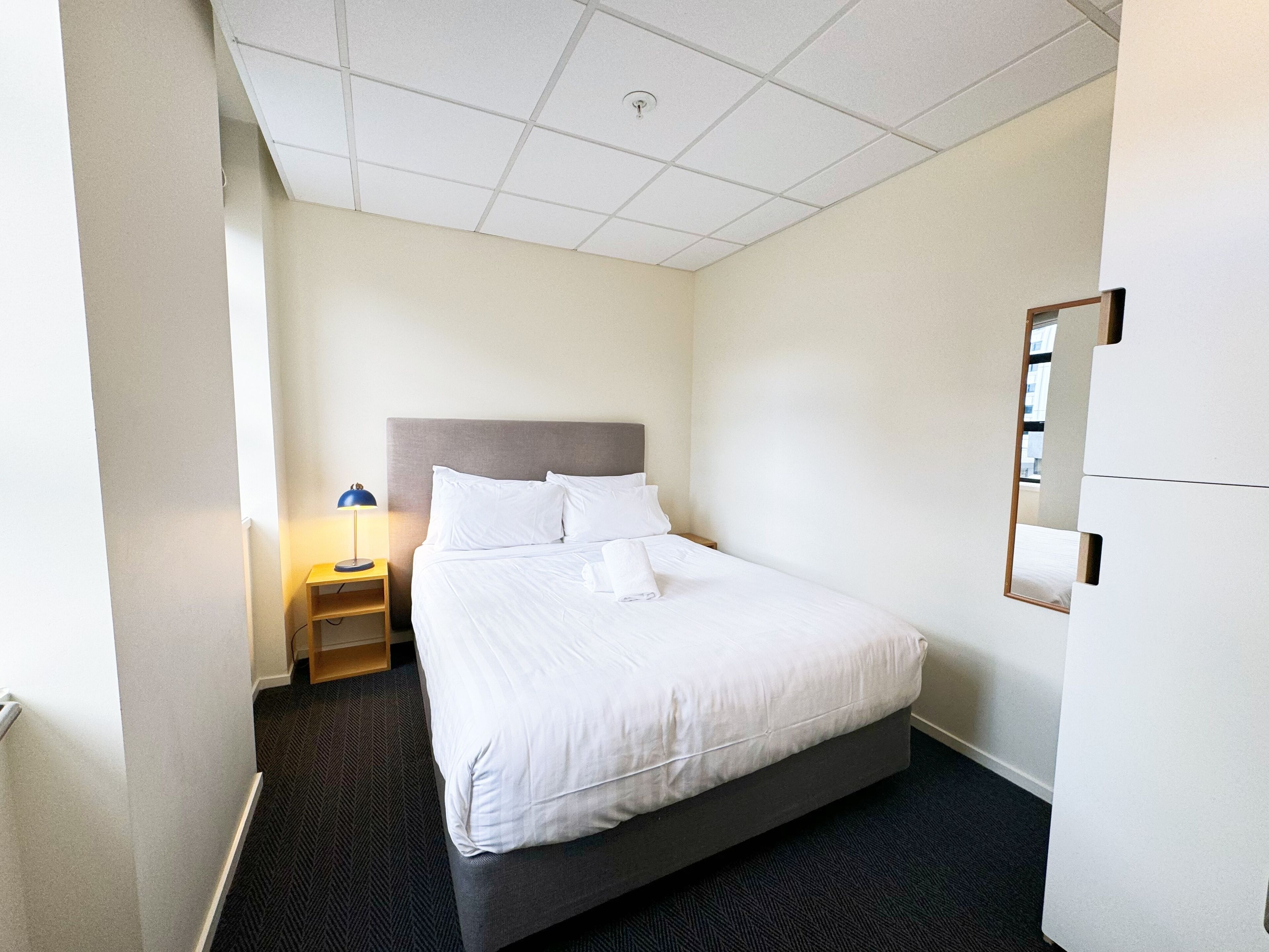 A hotel room with a large bed, a nightstand with a lamp, and a mirror on the wall.
