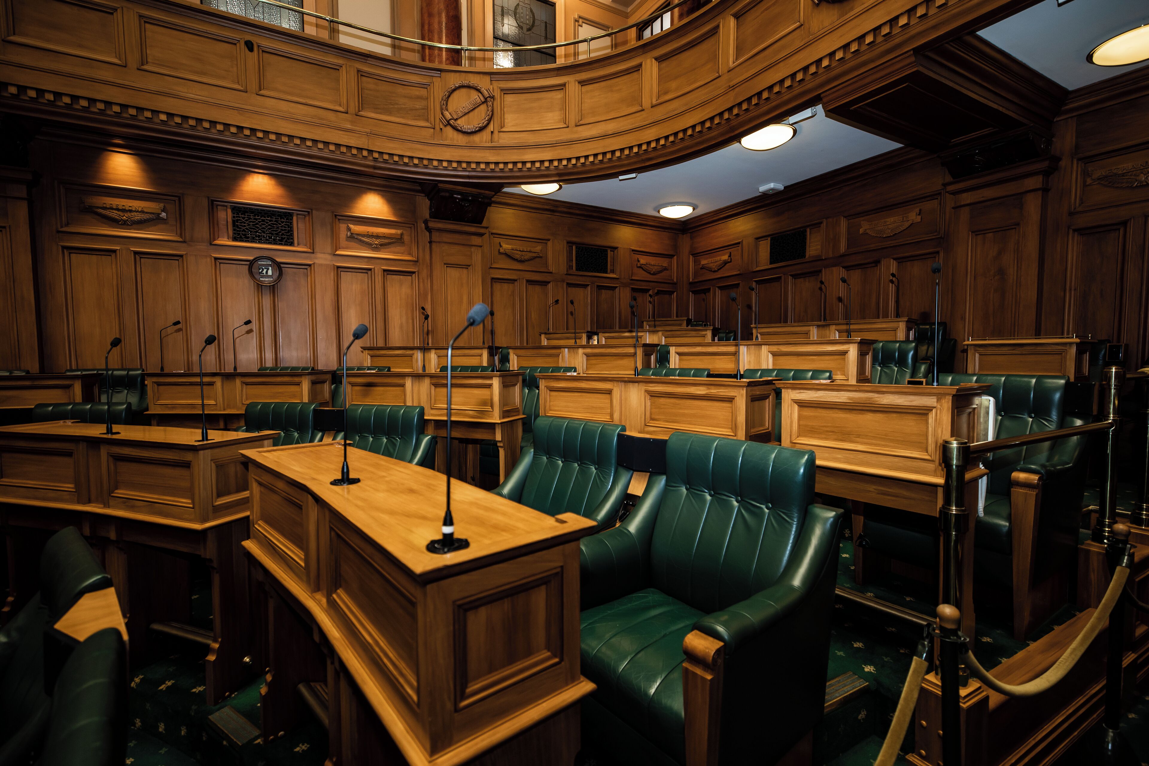 A formal meeting room with wooden paneling, green leather chairs, and a central podium with microphones.