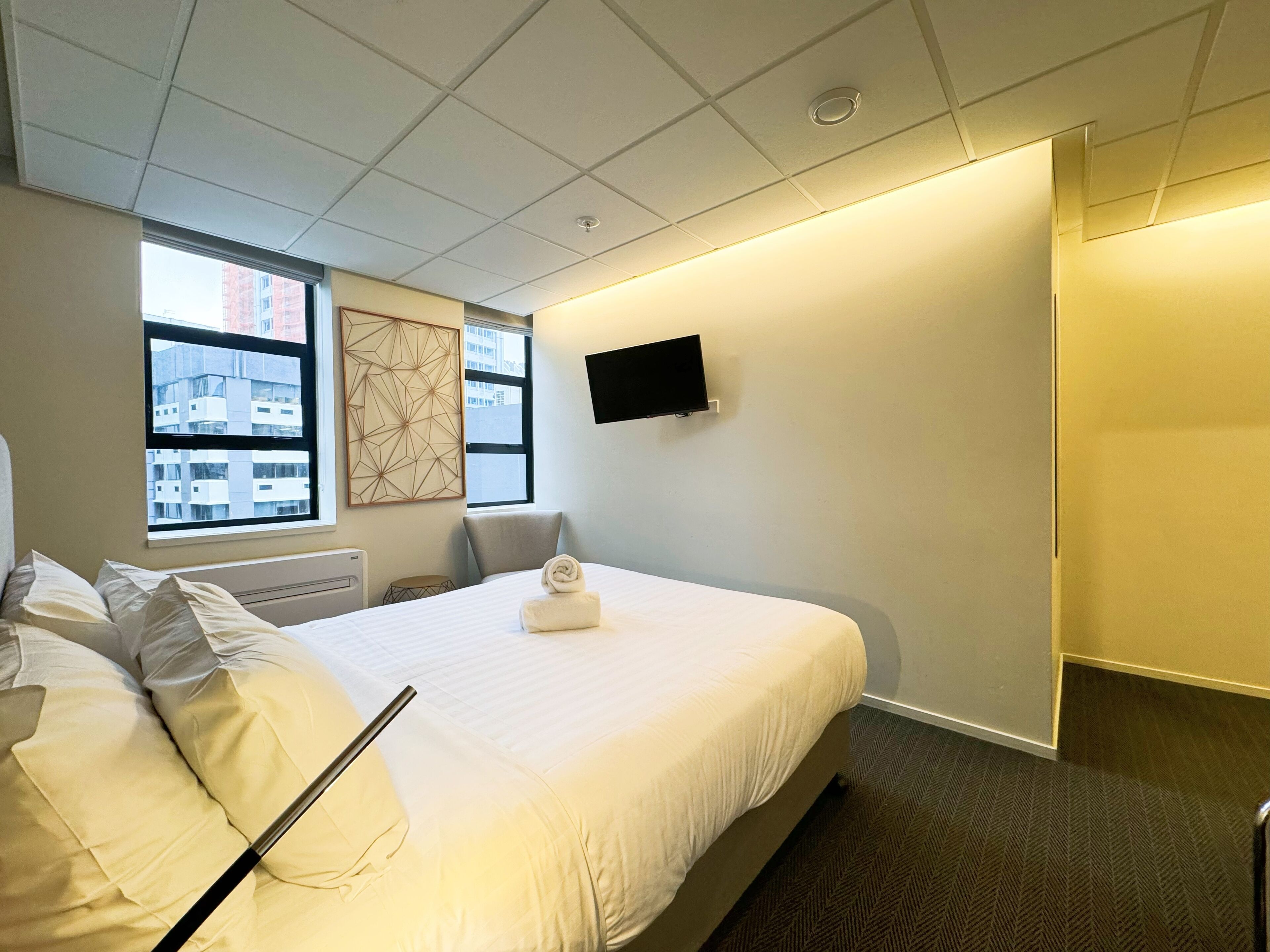 Habitación de hotel con una cama grande, un televisor de pantalla plana en la pared, una ventana con vista a la ciudad y una puerta a la derecha.