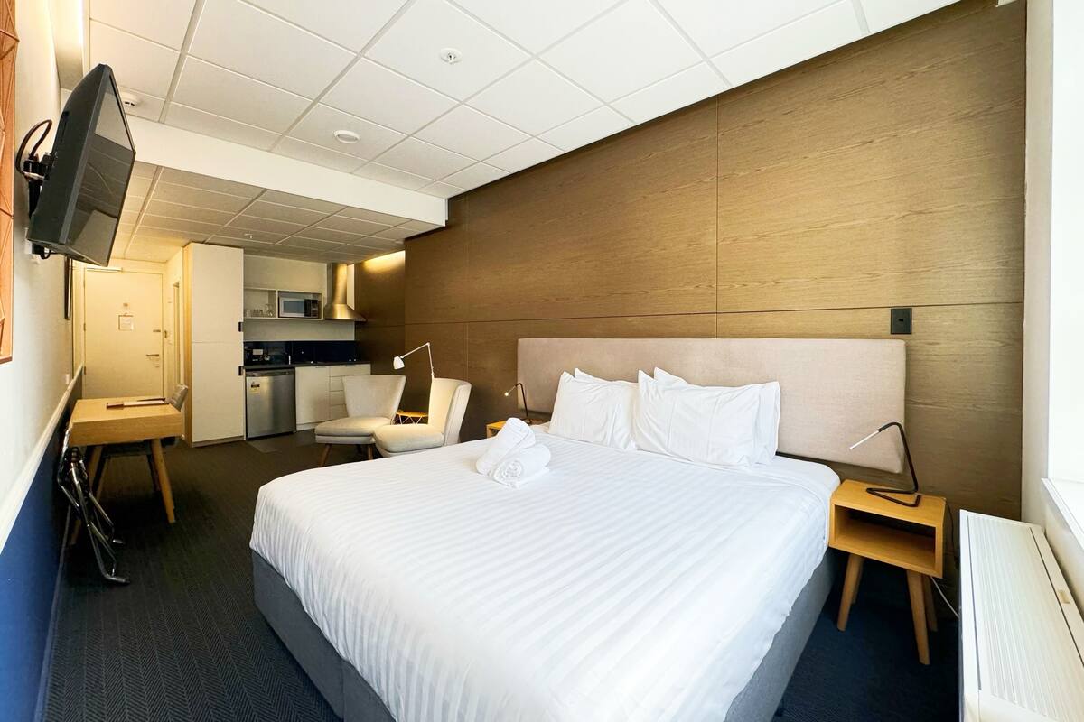 Habitación de hotel con una cama grande, un escritorio, una silla, un televisor y una kitchenette al fondo.