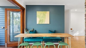 A dining area with a wooden table and green stools, a teal sofa, and a large abstract painting on the wall. - Cottage on Lorien Avenue (Coolum Beach)