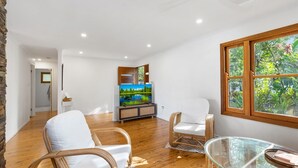 A spacious living room with a wooden floor, white furniture, a glass table, and a large window with a view of greenery. - Cottage on Lorien Avenue (Coolum Beach)