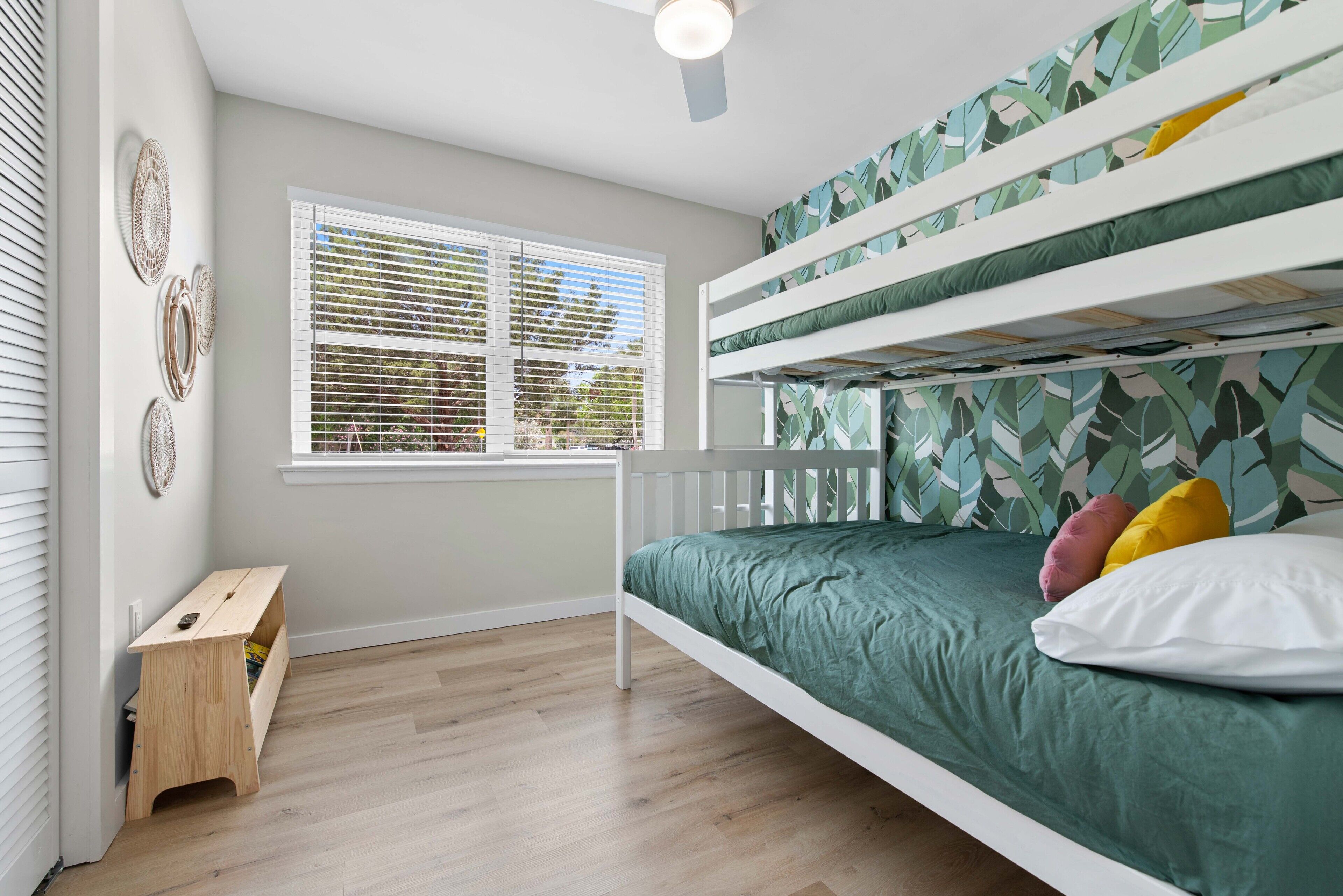 Una cama litera con un colchón verde y varias almohadas, un pequeño banco de madera y una ventana con persianas.