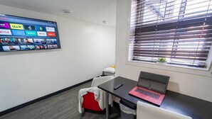 Ein modernes Zimmer mit Schreibtisch, Laptop, an der Wand montiertem Fernseher, der verschiedene Streaming-Dienste anzeigt, Fenster mit Jalousien und einer kleinen Pflanze am Fensterbrett.