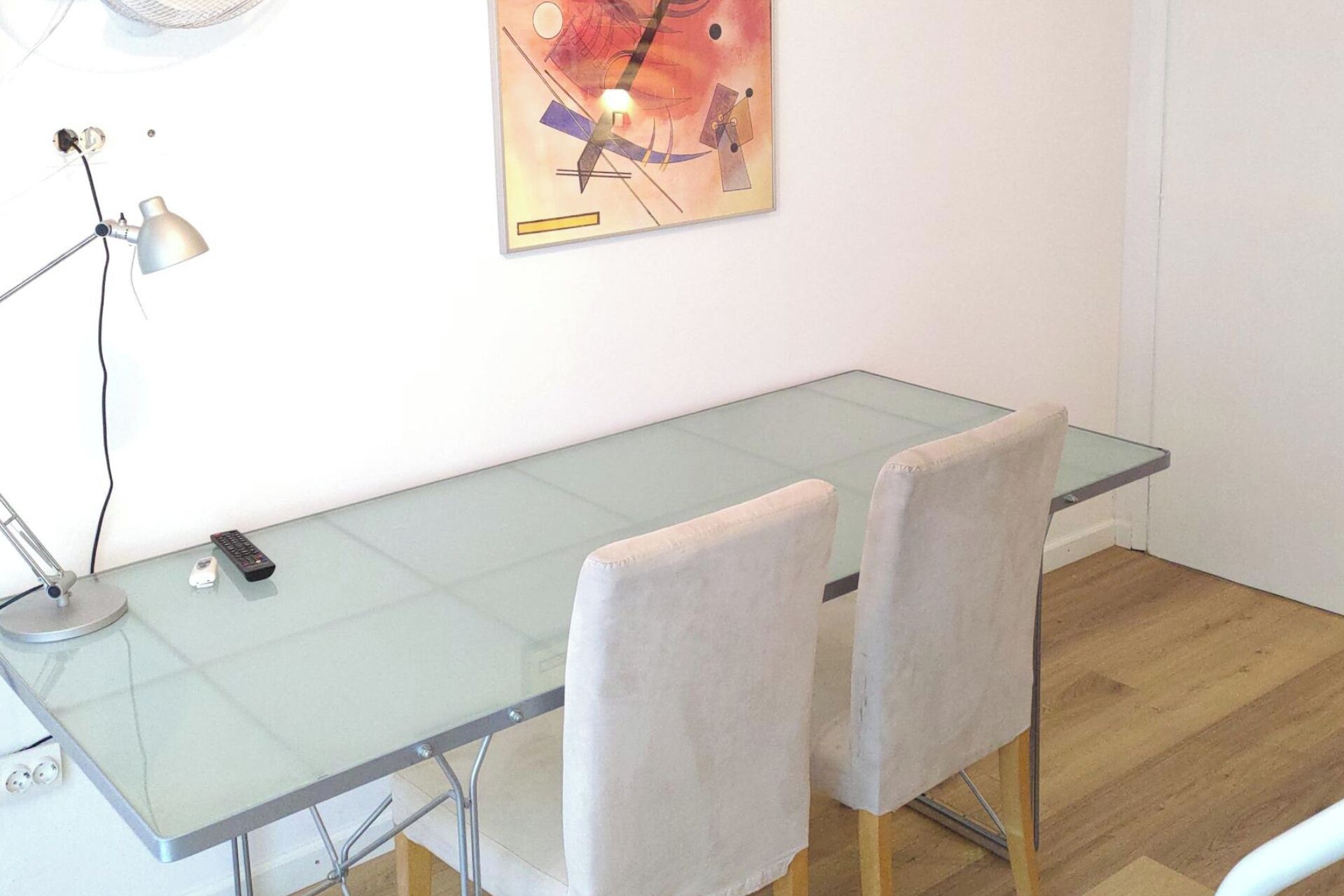 Ein Zimmer mit einem Glastisch, zwei Stühlen, einer Schreibtischlampe, einer Fernbedienung und einem an der Wand hängenden abstrakten Gemälde.