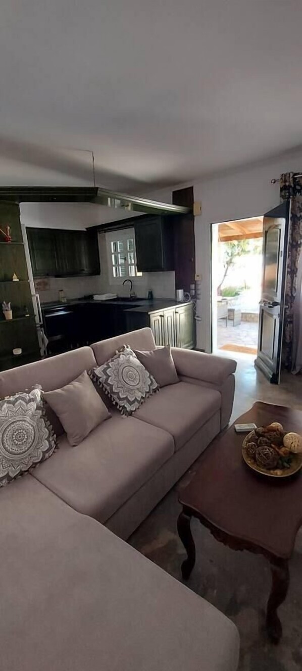 A living room with a beige sofa, patterned cushions, and a wooden coffee table with a bowl of bread. A kitchen is visible in the background with dark cabinets and a window.