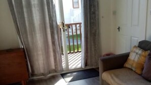 A room with a wooden floor, a sofa with a plaid cover, a wooden side table, and a sliding glass door leading to an outdoor area with a railing. - Chalet Retreat in Mablethorpe (Mablethorpe)