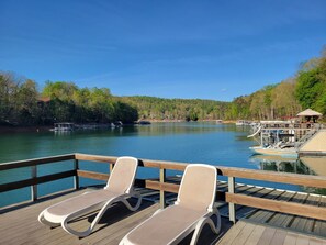 Une terrasse en bois avec deux chaises longues donnant sur un lac paisible, entourée d’arbres et bordée d’un quai où sont amarrés des bateaux.