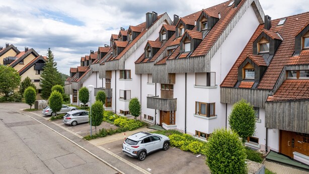 Une rangée d’immeubles résidentiels modernes, dotés de toits inclinés et de balcons, un parking et des espaces verts aménagés.