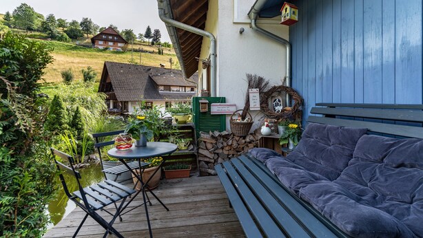 Um deck de madeira com um banco e mesa, rodeado por vegetação e vista para uma casa.