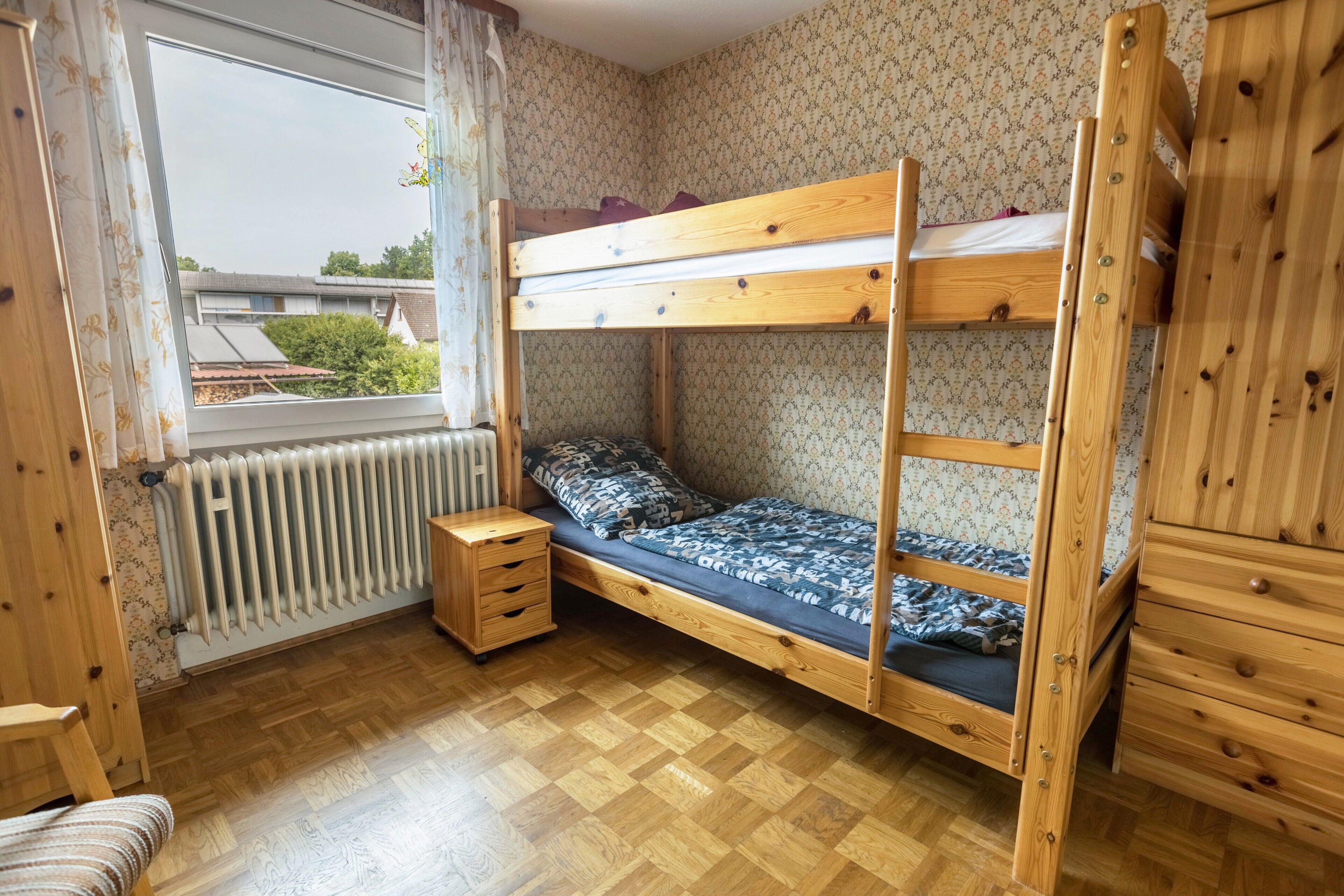 Ein Schlafraum mit Etagenbetten, Holzboden, Heizkörper, einem Fenster mit Vorhängen und einem Holzschrank.