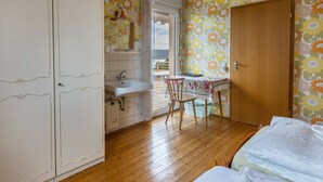 Una camera da letto con pavimento in legno, un letto, un tavolino con una sedia, un bagno con un lavandino e una porta che dà su un balcone.