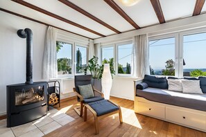 A cozy room with a wood-burning stove, a comfortable sofa, a chair, and large windows offering a view of the outdoors.