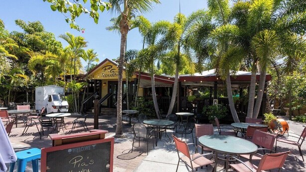 Un espace extérieur aménagé en terrasse, avec des tables et des chaises, entouré de palmiers et d’un bâtiment sur lequel on peut lire l’inscription « Pia’s Veranda ».