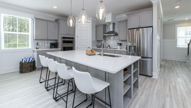 Una cocina moderna con una isla grande, taburetes blancos tipo bar, electrodomésticos de acero inoxidable e iluminación colgante.