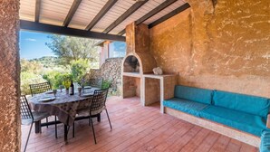 Patio esterno con parete in pietra, tetto in legno, forno a pietra, tavolo apparecchiato per mangiare e panca imbottita blu.