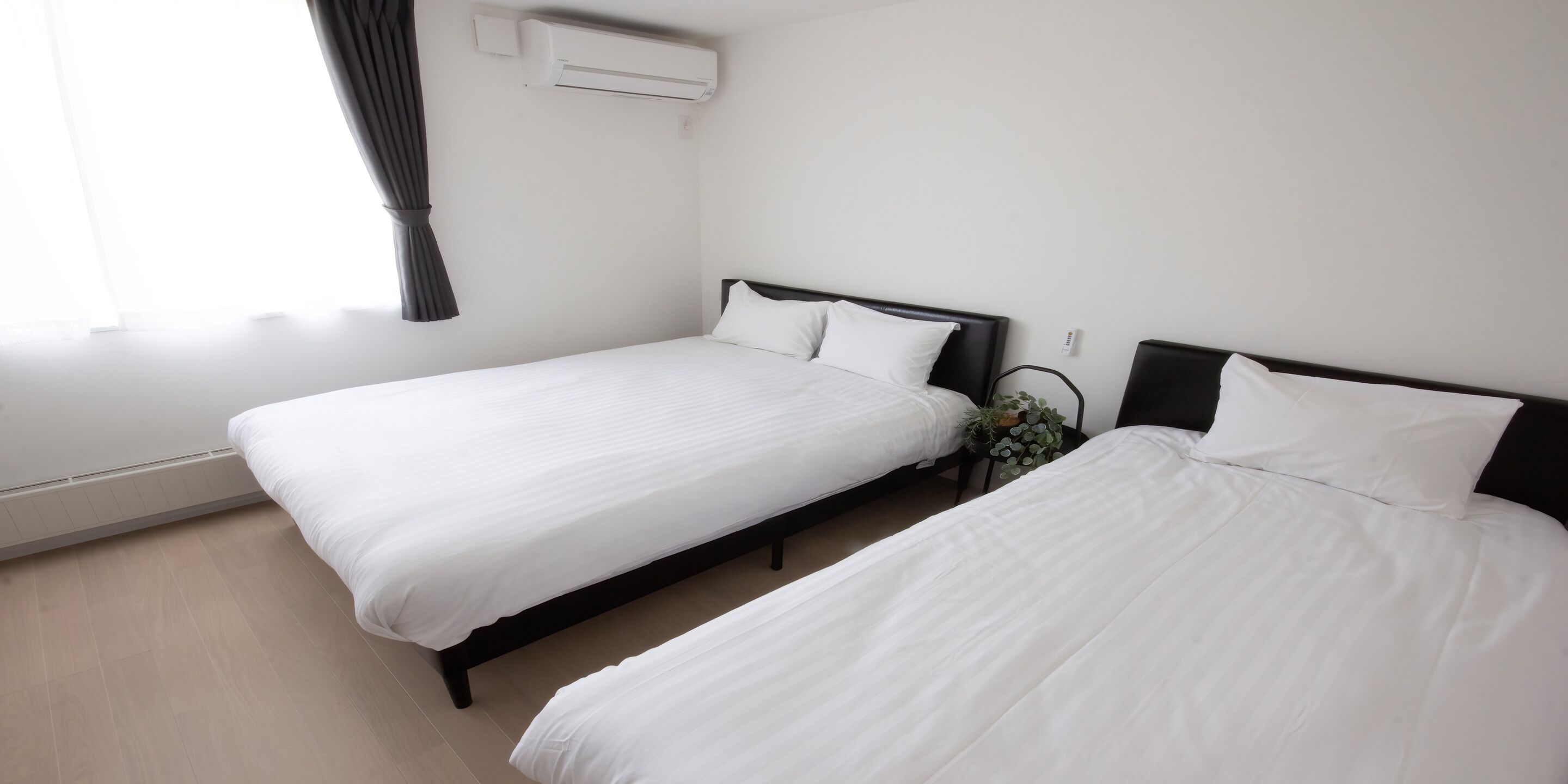 Dos camas individuales con ropa de cama blanca, una ventana con cortinas, un aire acondicionado de pared y una pequeña planta en maceta.