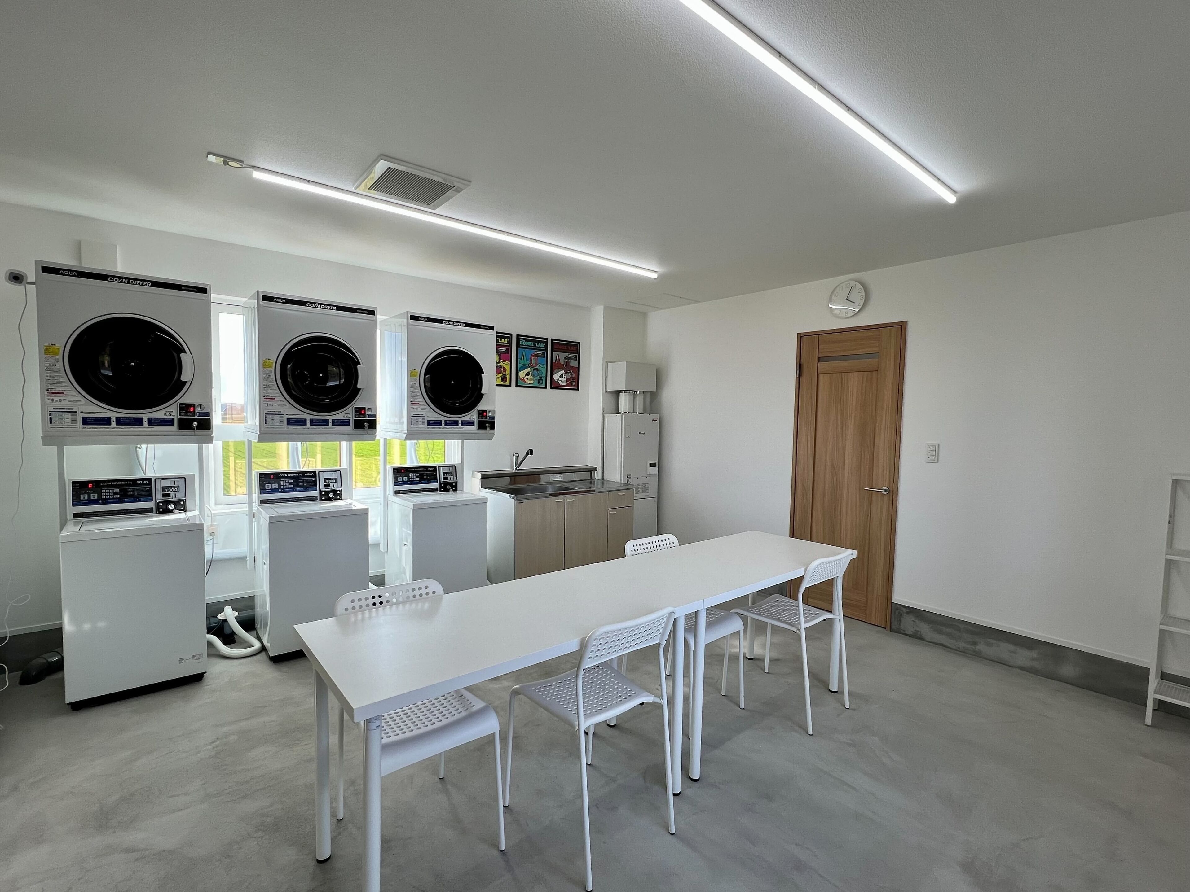 A laundry room with three washing machines, a sink, a table with chairs, and a wooden door.