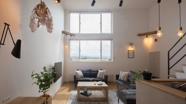 Una sala de estar moderna con un ventanal grande, un sofá negro, una mesa de centro de madera y una escalera.