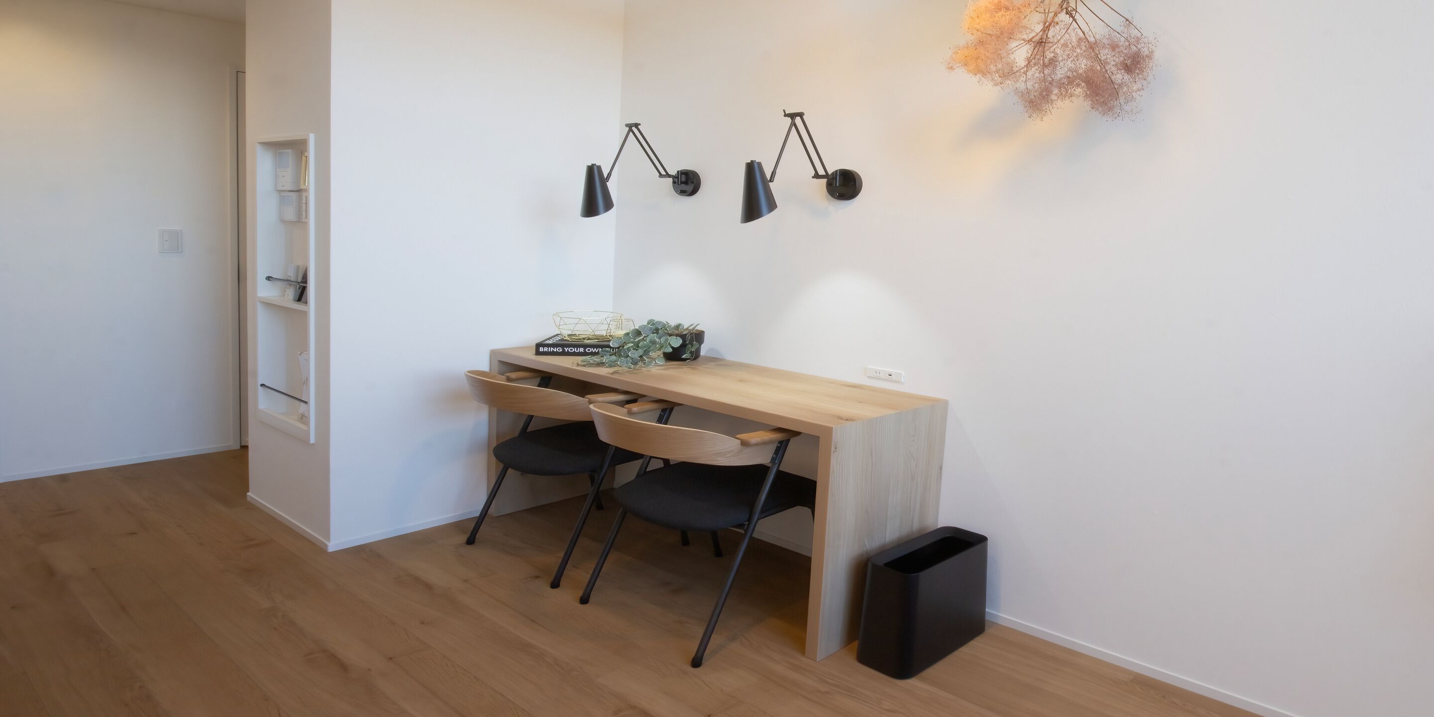 A small, modern room with a wooden desk, two chairs, a black trash bin, and a wall-mounted lamp.