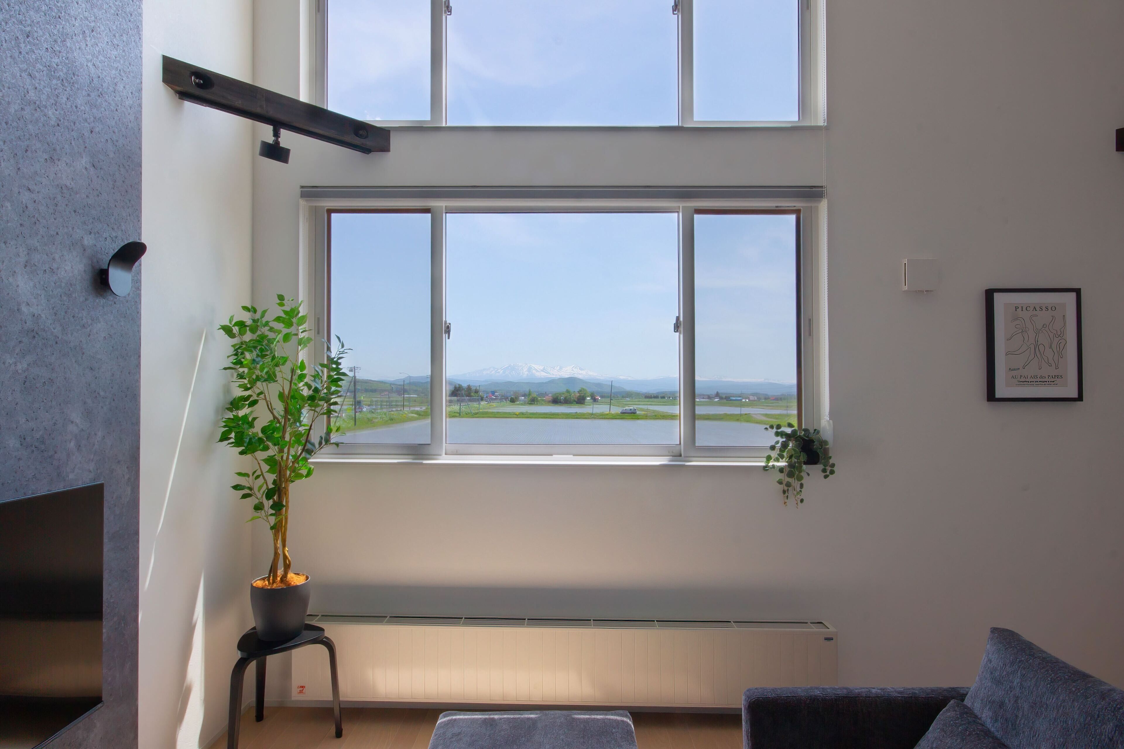 A modern living room with a large window, a potted plant, a flat-screen TV, and a framed artwork on the wall.