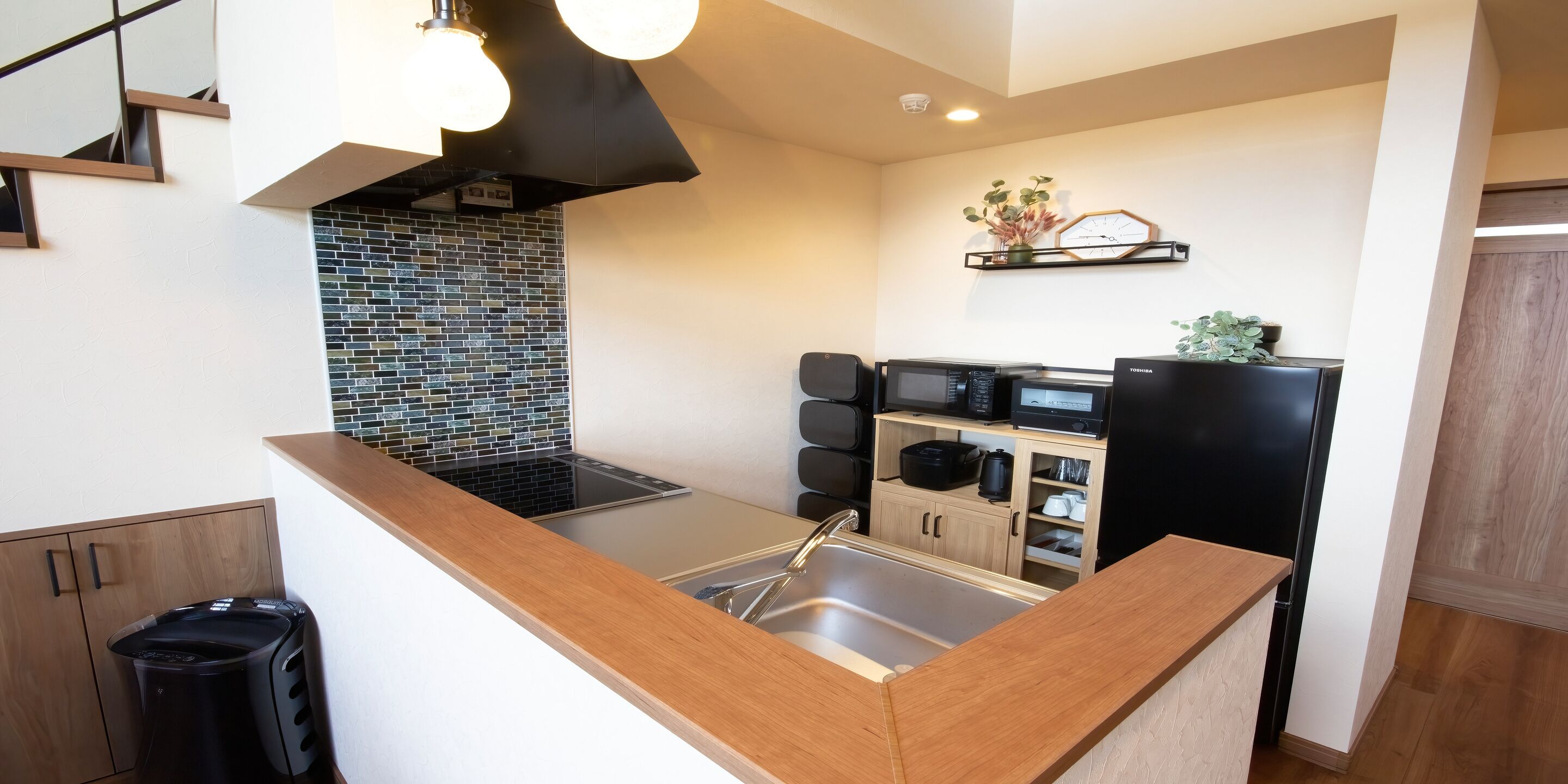 A modern kitchen with a black stove, stainless steel sink, and wooden countertop. There is a black trash bin and a microwave on a shelf.