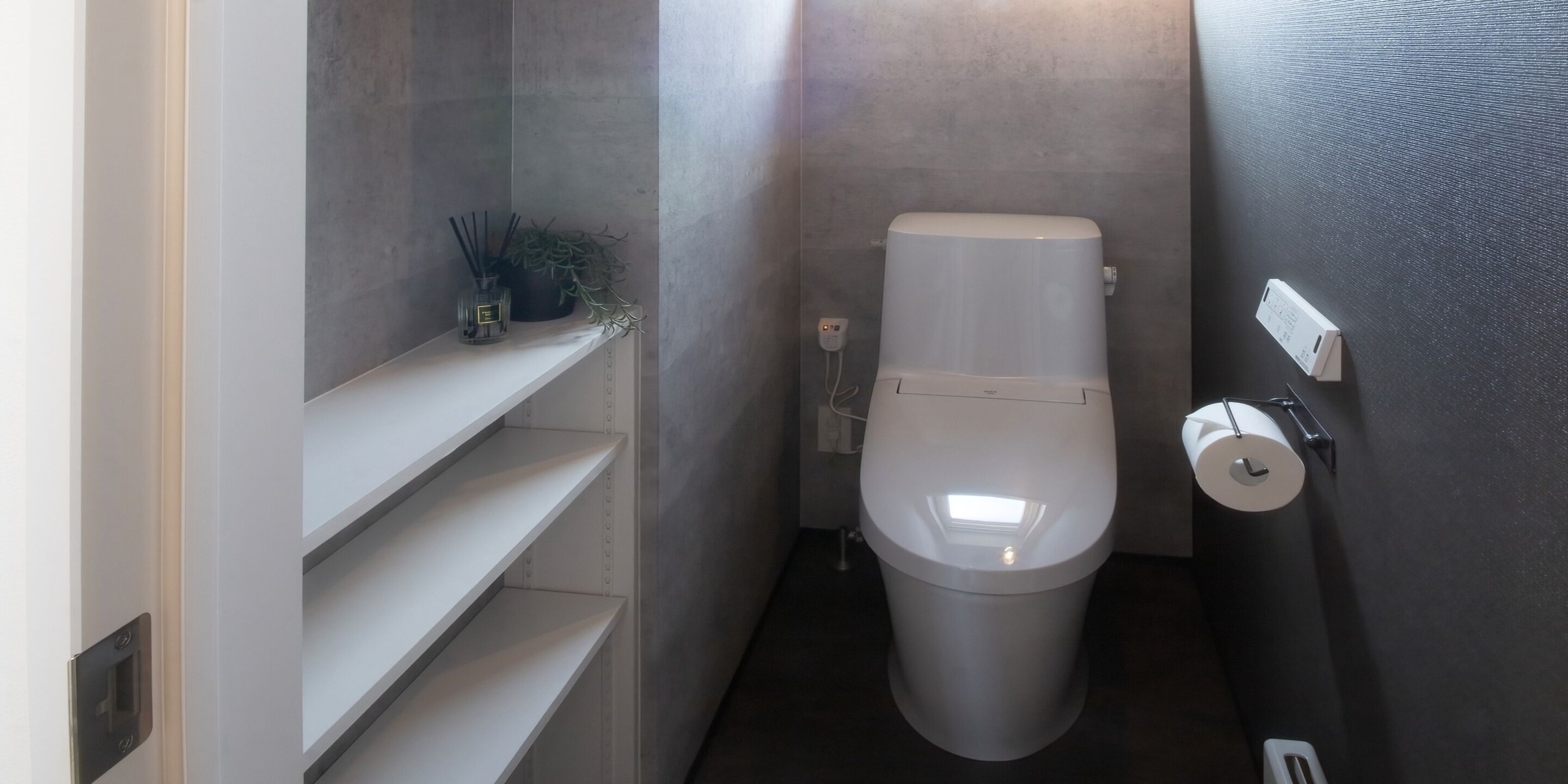 A compact bathroom with a toilet, a shelf with a plant and a glass jar, and a toilet paper holder on the wall.