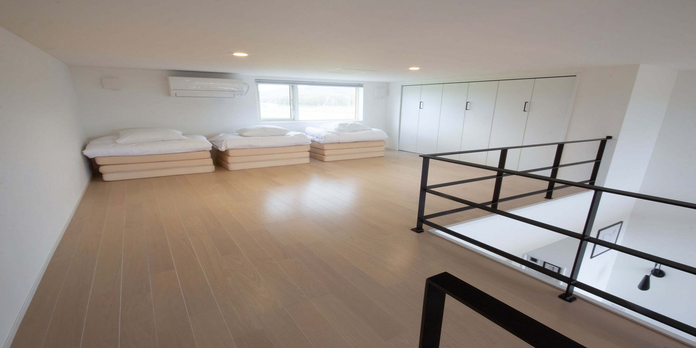 A spacious room with a wooden floor, a bed with white bedding, a window, and a wardrobe.