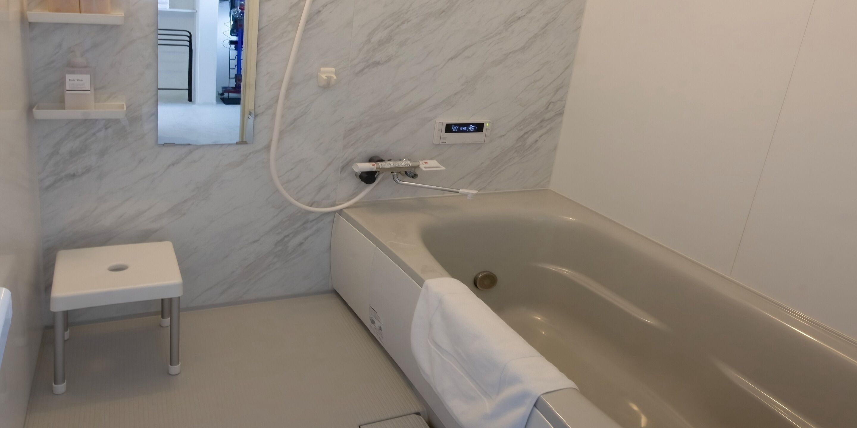 Une salle de bain moderne avec un mur en marbre, un lavabo sur colonne blanc, une baignoire avec une serviette et un thermostat numérique.
