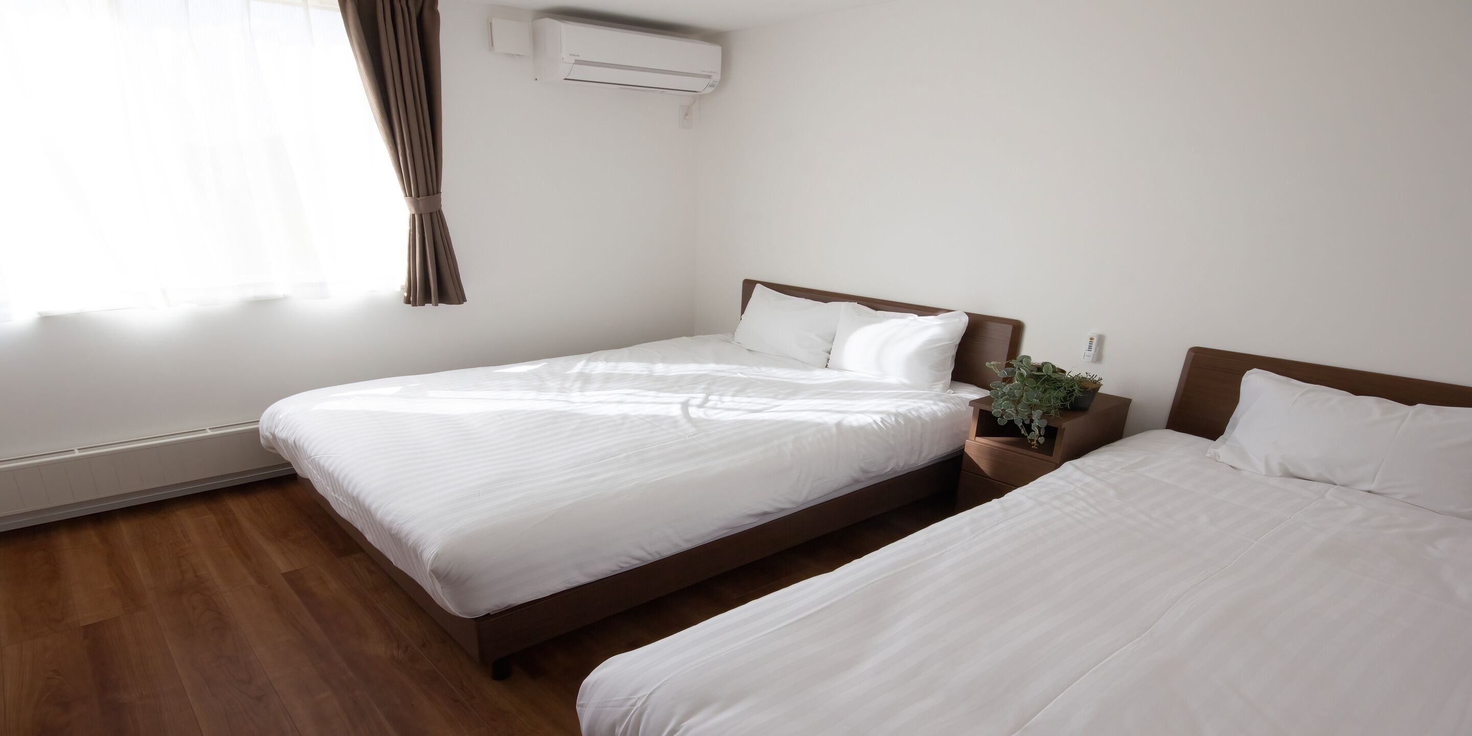 A hotel room with two single beds, a wooden floor, a window with curtains, and an air conditioning unit.