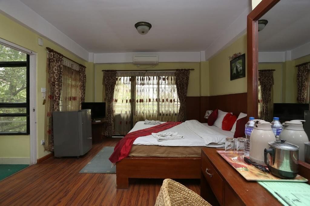 A hotel room with a wooden bed, a desk with a kettle, a refrigerator, and a window with curtains.
