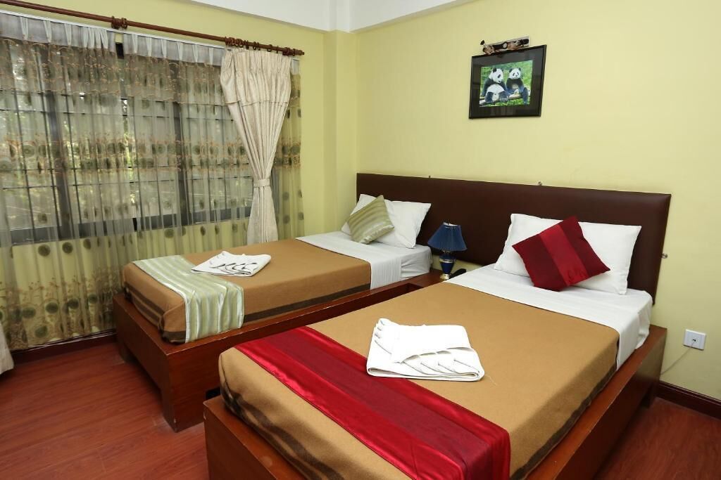 A hotel room with two beds, a window with curtains, a framed picture on the wall, and a red and white pillow on one bed.