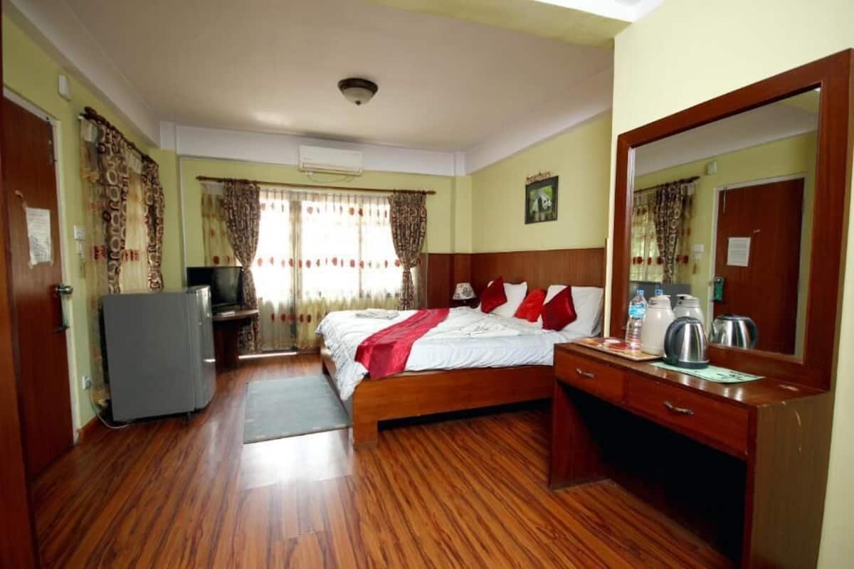 Une chambre d’hôtel avec un plancher en bois, un lit avec une literie rouge et blanche, un bureau avec un miroir, une télévision et un réfrigérateur.