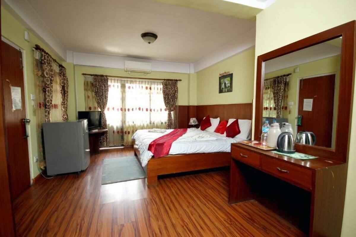 Une chambre d’hôtel avec un plancher en bois, un lit avec des oreillers rouges, un bureau avec un miroir, une télévision et un réfrigérateur.