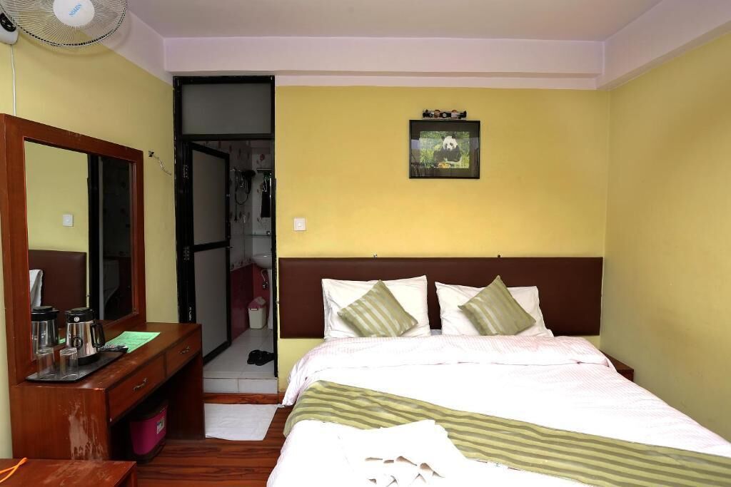 A hotel room with a bed, a wooden desk, a mirror, a fan, and a framed picture on the wall.