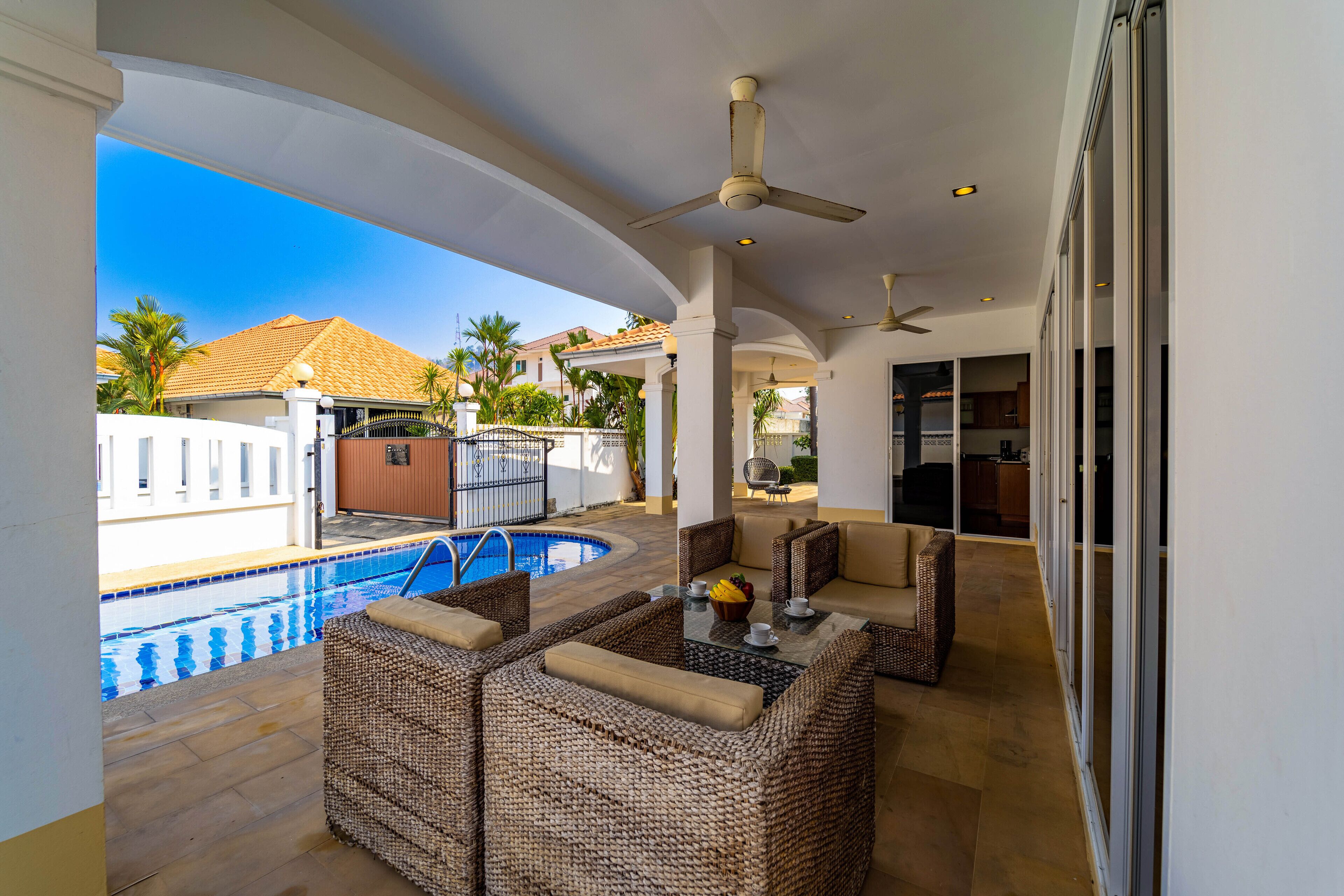 Un espace extérieur abrité, aménagé avec des meubles en osier, une piscine et offrant une vue sur une maison dont le toit est recouvert de tuiles.
