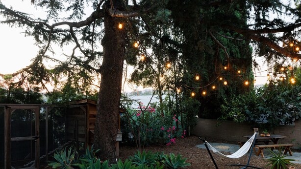 Un hamac suspendu sous des guirlandes lumineuses, une table en bois et un jardin avec des plantes et un arbre.