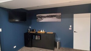 Habitació amb parets blaves fosques, un televisor pantalla plana fixat a la paret, una cuineta petita amb torradora, tetera i pots, i una porta amb marc blanc.