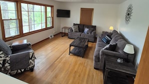 A spacious living room with hardwood flooring, a grey sofa set, a coffee table, and a TV mounted on the wall.