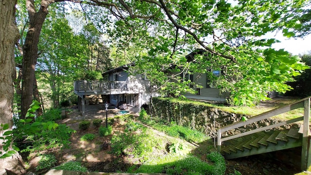 A two-story house with a balcony, surrounded by lush greenery and a stone wall.