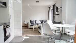 Una cocina moderna con armarios blancos y horno empotrado, un comedor con una mesa redonda y sillas blancas, y una sala de estar con sofá y mesa de centro.