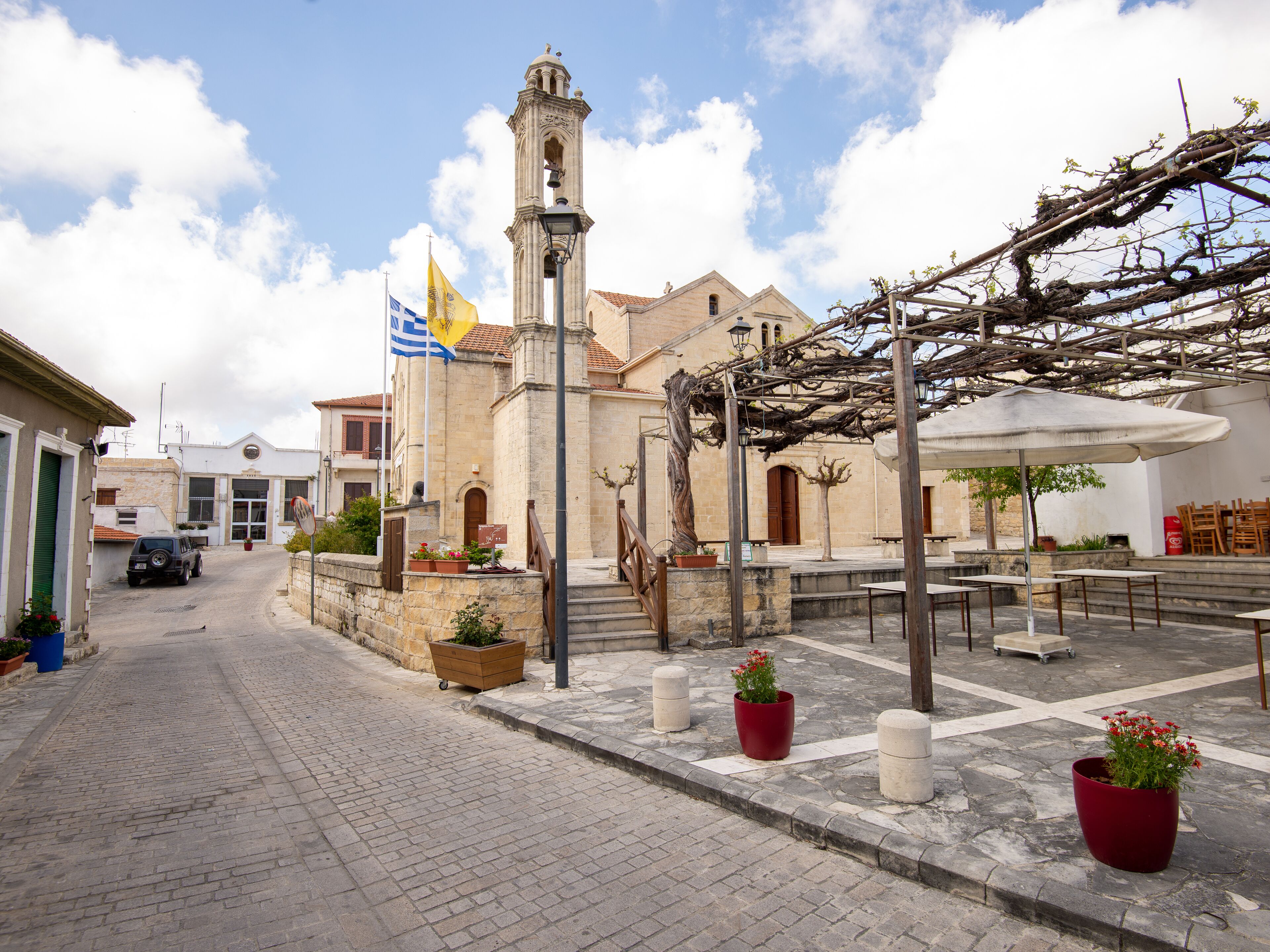 A cobblestone street with a historic building featuring a bell tower, outdoor seating, and potted plants.