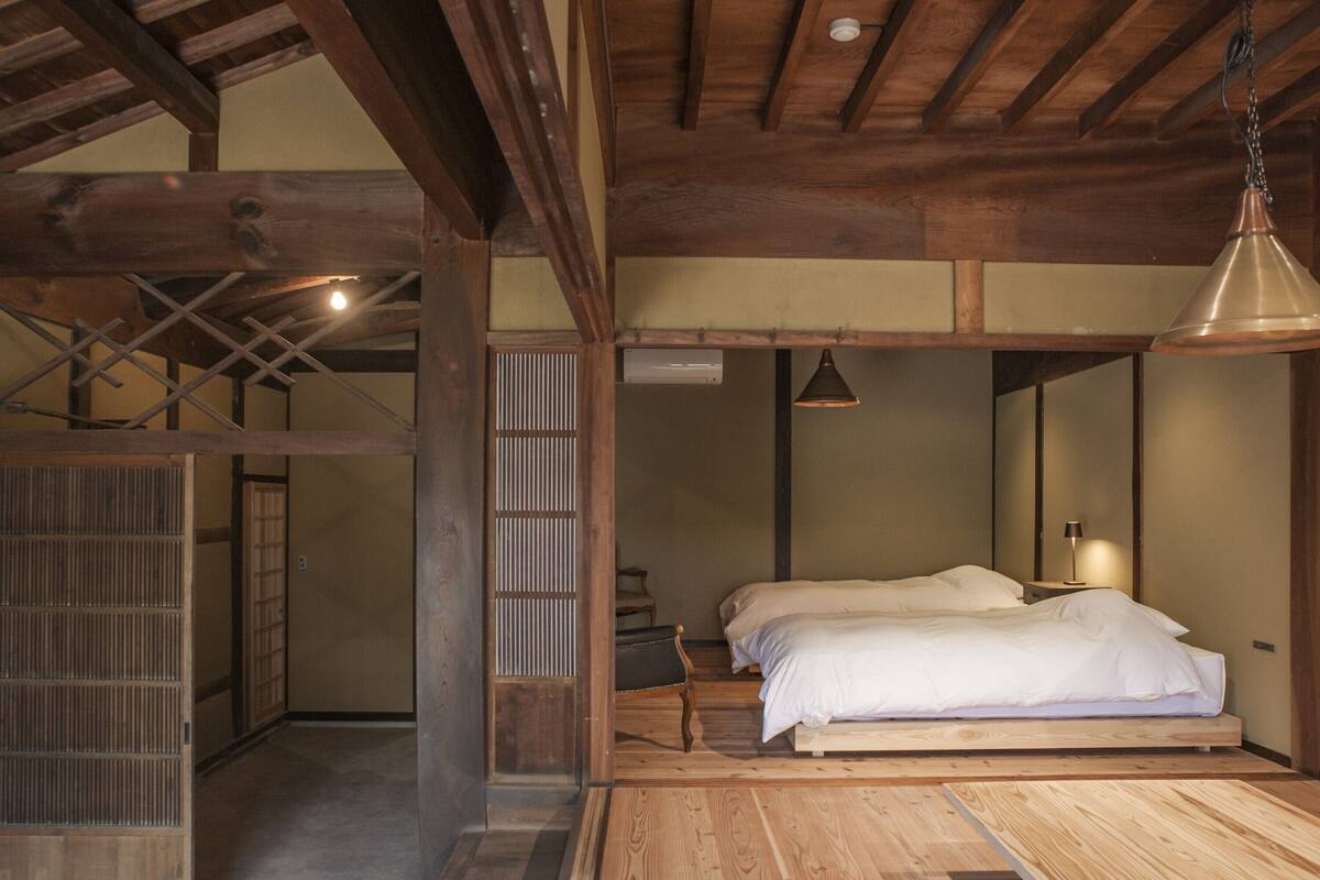 A traditional Japanese-style room with a wooden floor, a bed with white bedding, a hanging lamp, and a sliding door.