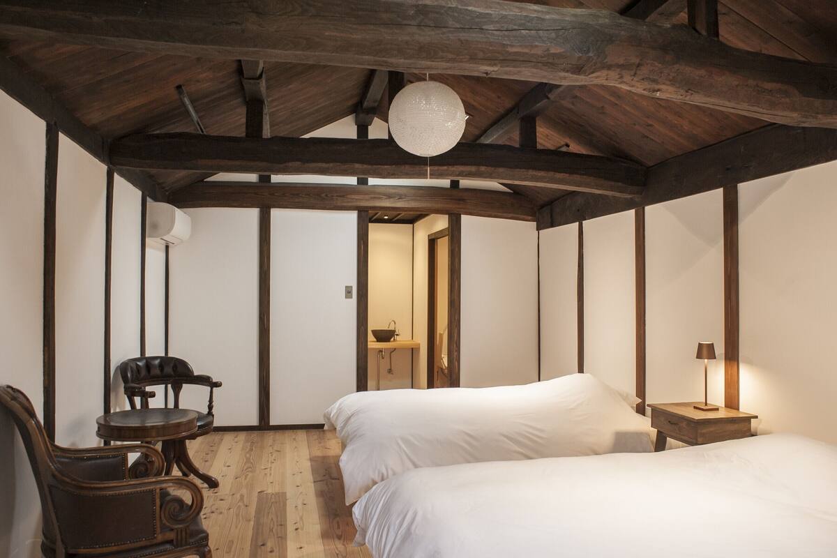A bedroom with two beds, a wooden chair, a small table, a bedside lamp, and a hanging light fixture.