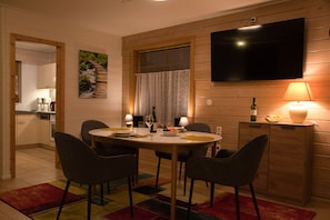 Un coin repas avec une table ronde, des chaises, un téléviseur fixé au mur, une lampe et une bouteille de vin.