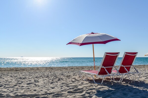 A beach scene with two red and white beach chairs and a matching umbrella under a clear sky.
